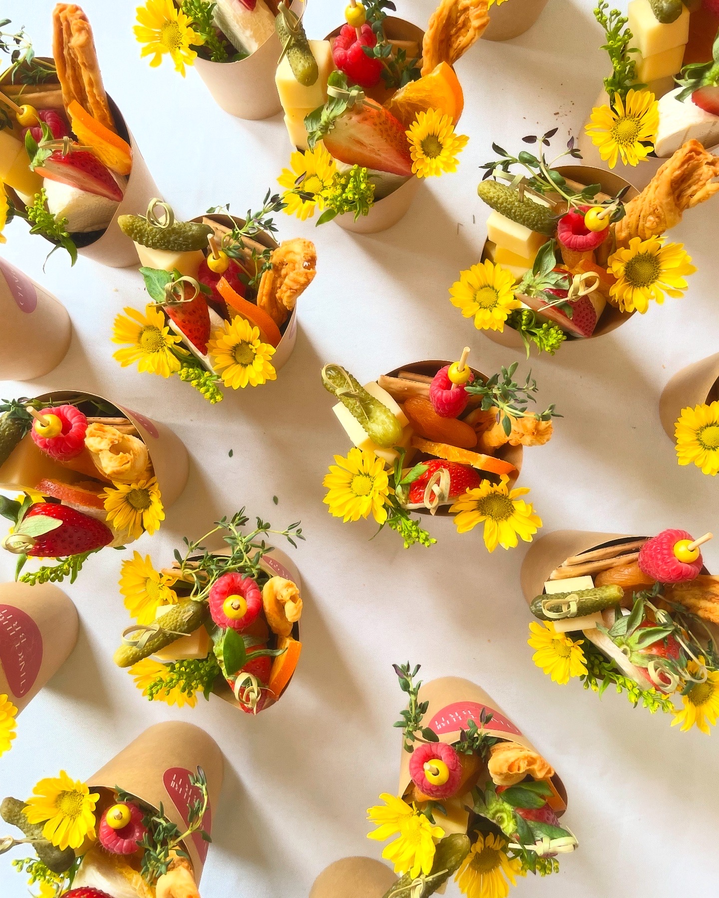 Charcuterie cups with three cheeses, salami, cornichons, fresh fruit, roasted nuts, crackers, and a floral garnish, individually styled for guests